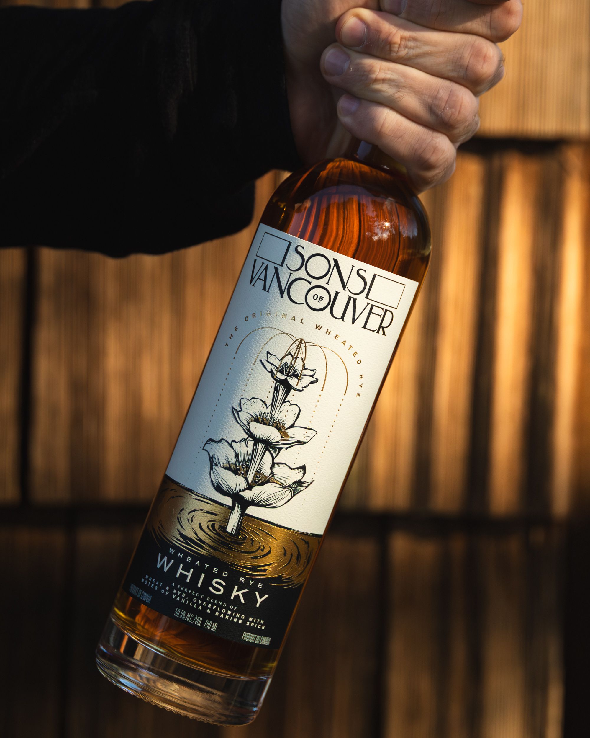 Hand gripping Sons of Vancouver Wheated Rye bottle against warm slatted wood