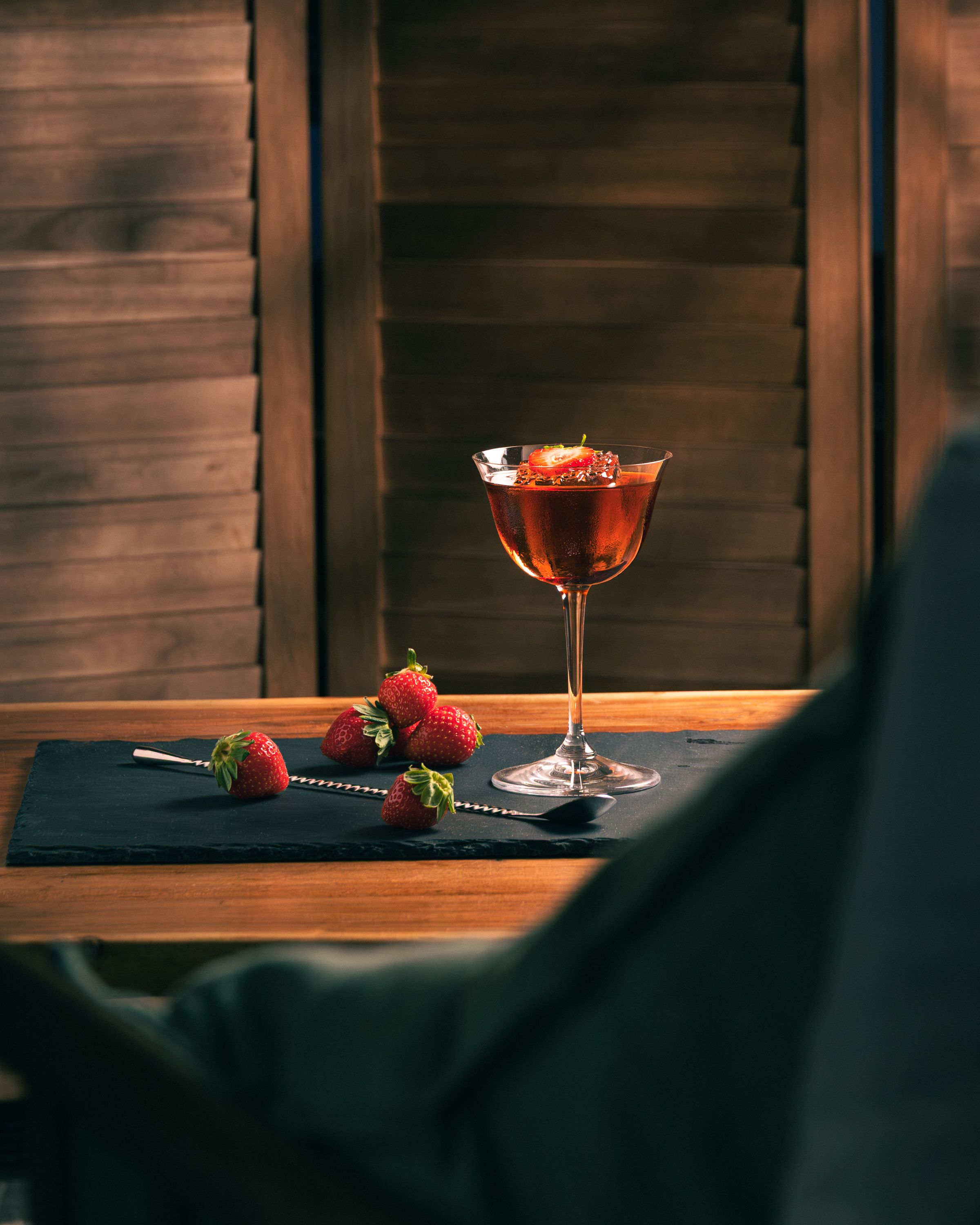Strawberry negroni in coupe with fresh strawberries and bar spoon on slate, wood shutters behind