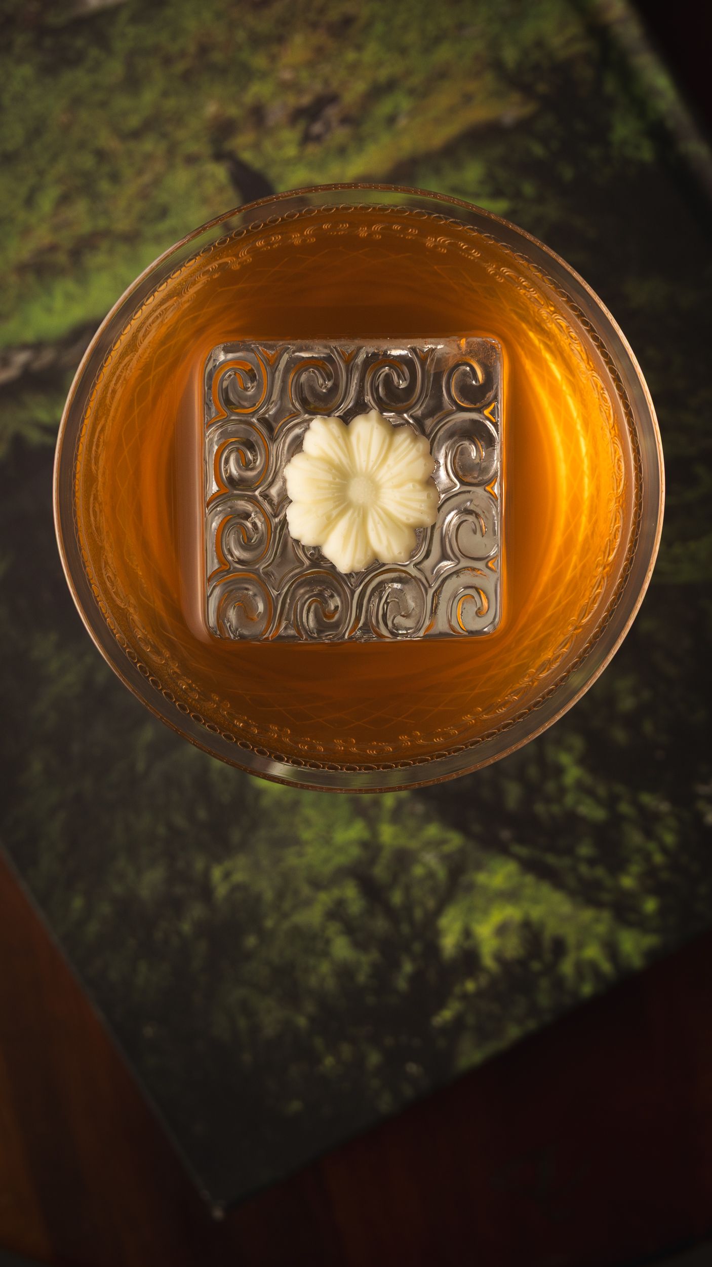Top-down whisky cocktail with a carved floral ice cube on a mossy green surface.