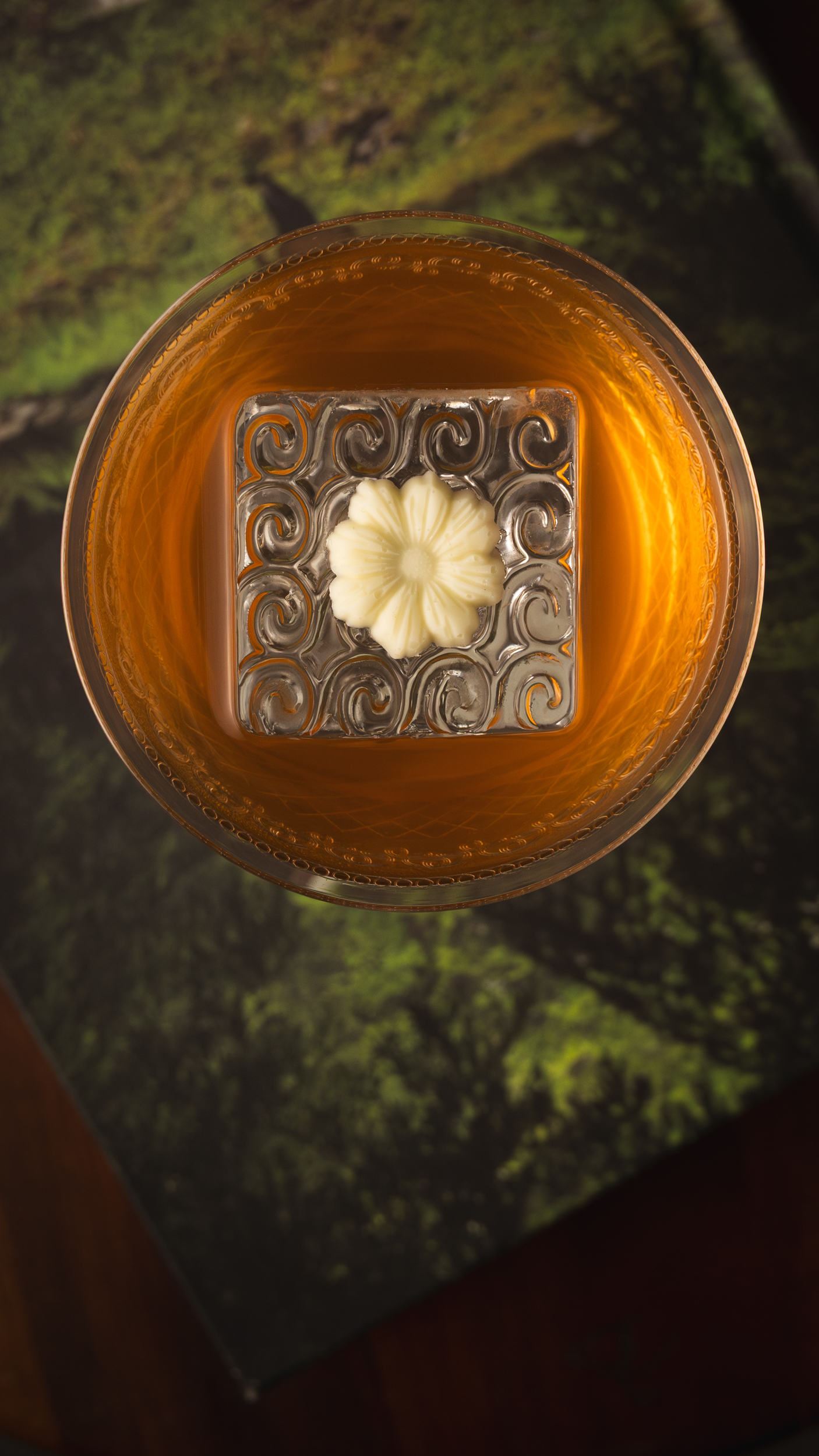 Top-down whisky cocktail with a carved floral ice cube on a mossy green surface.