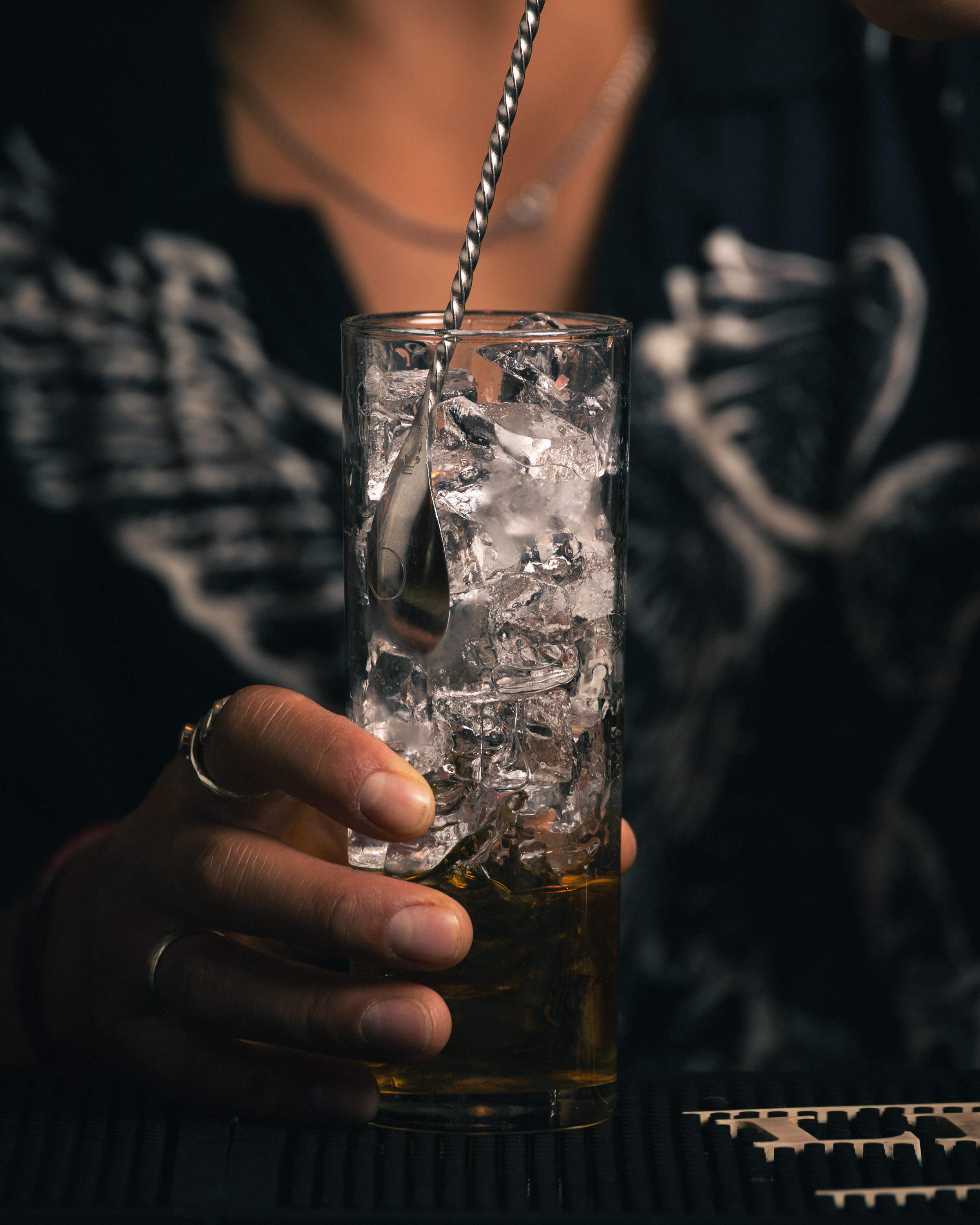 Person holding a whisky highball glass with ice being stirred at a bar