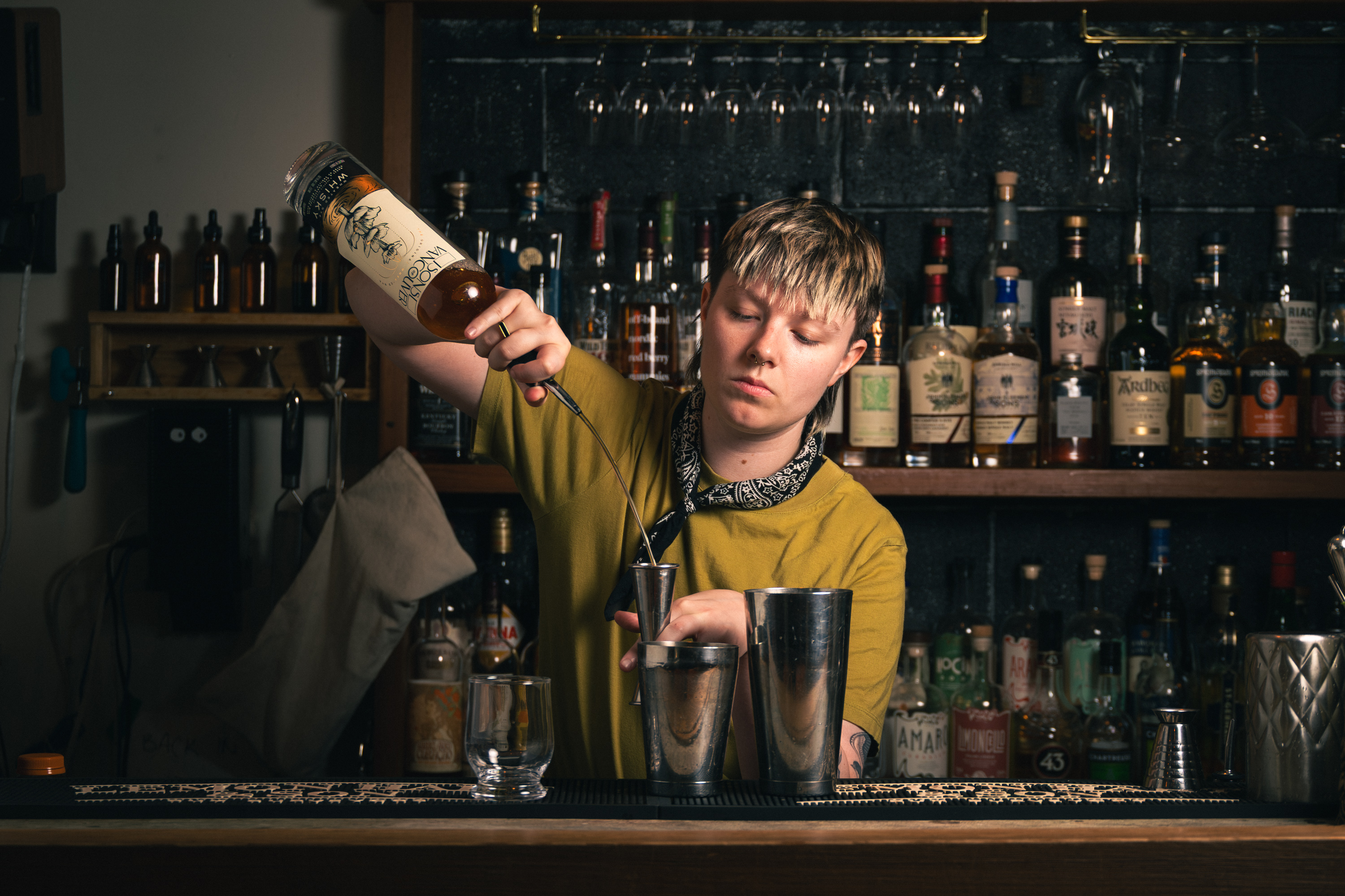Bartender measuring whisky into a jigger at a well-stocked bar