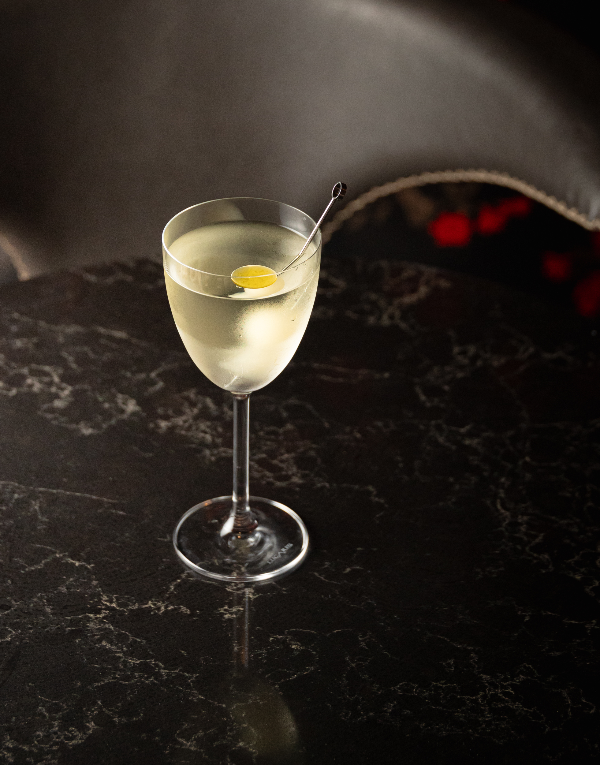 Pale martini with lemon twist on a pick on black marble, leather chair and red lights behind