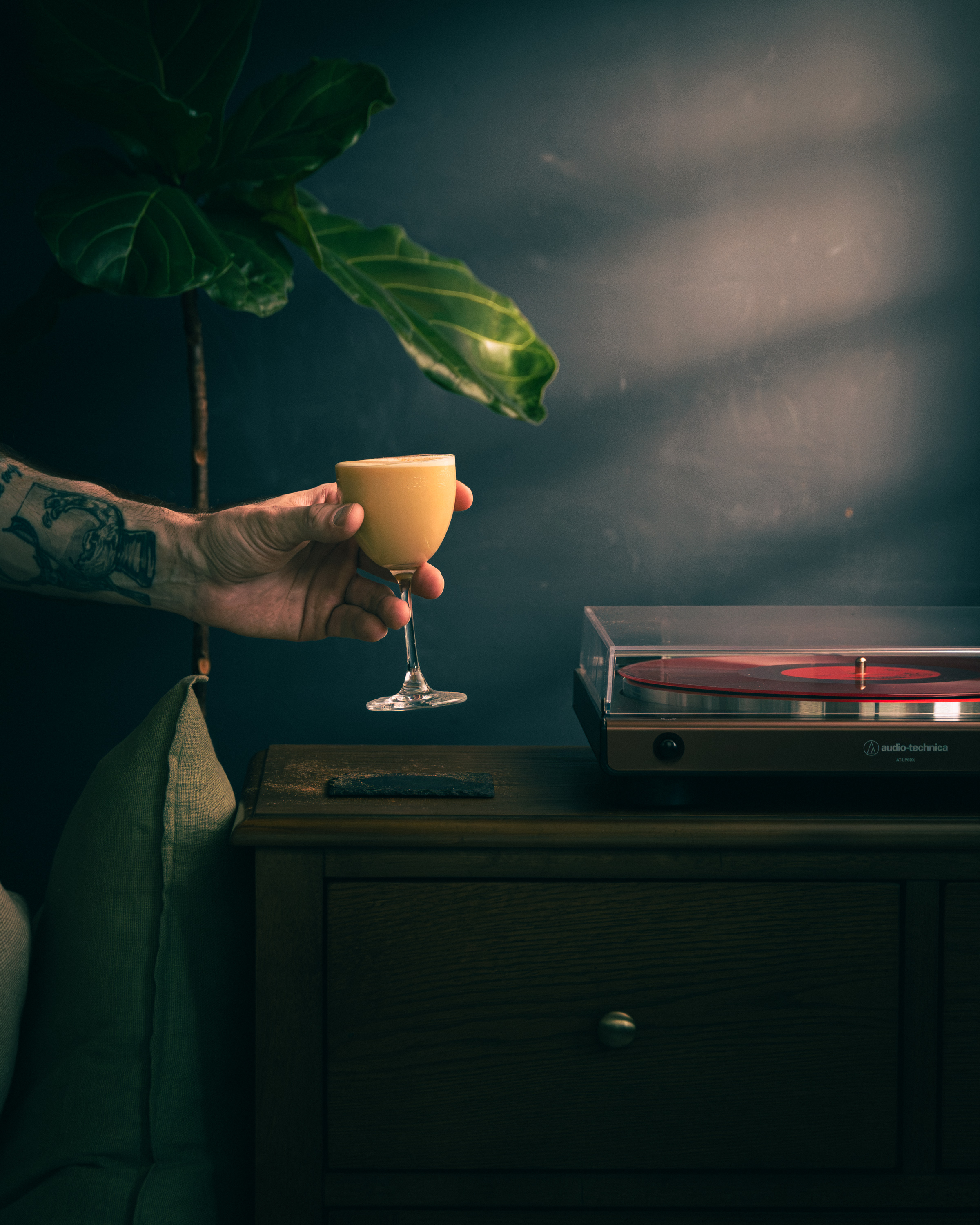 Tattooed hand holding a golden coffee flip in stemmed glass beside a red vinyl record player