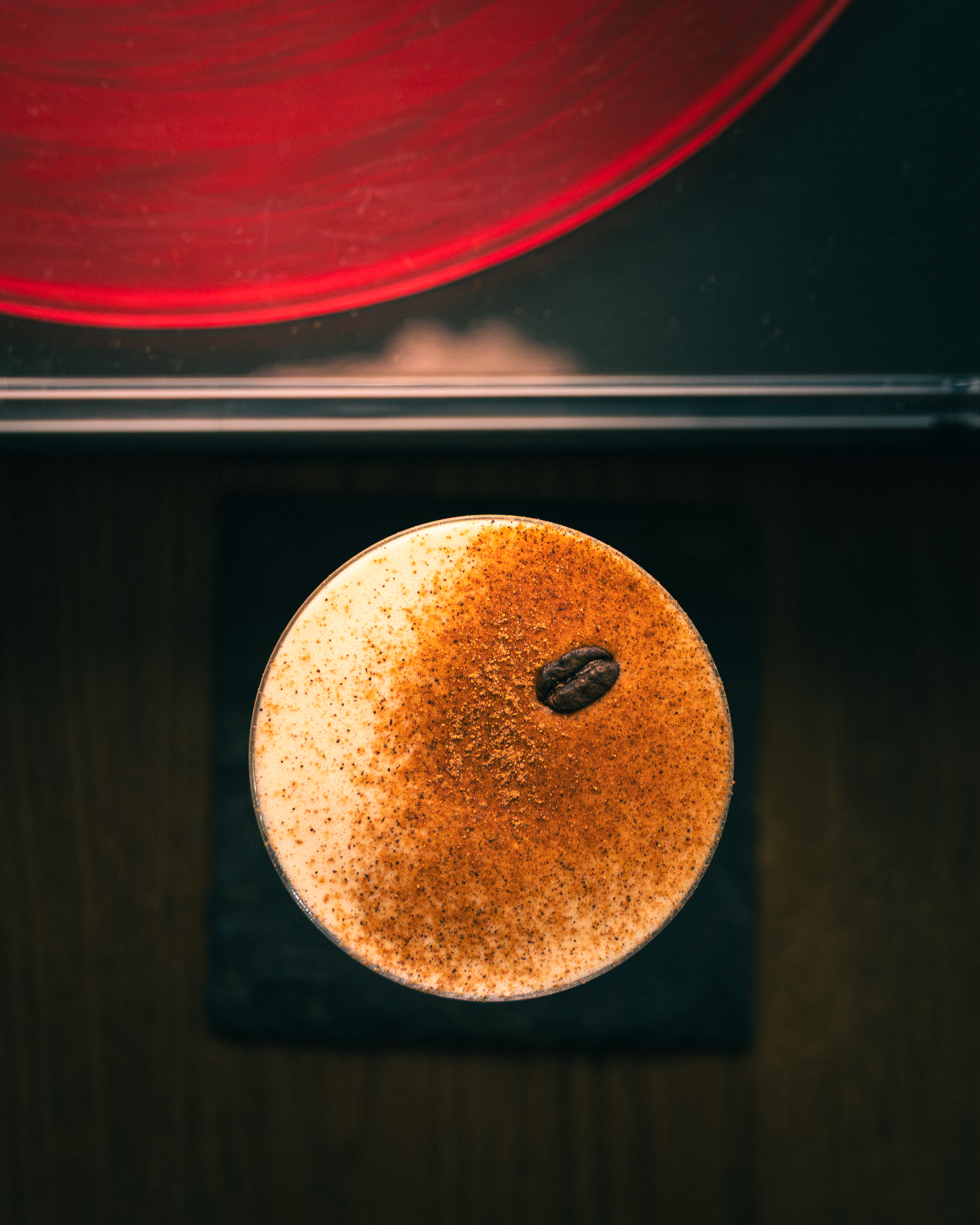 Top-down coffee flip with cinnamon-dusted foam and single coffee bean beside a red vinyl record