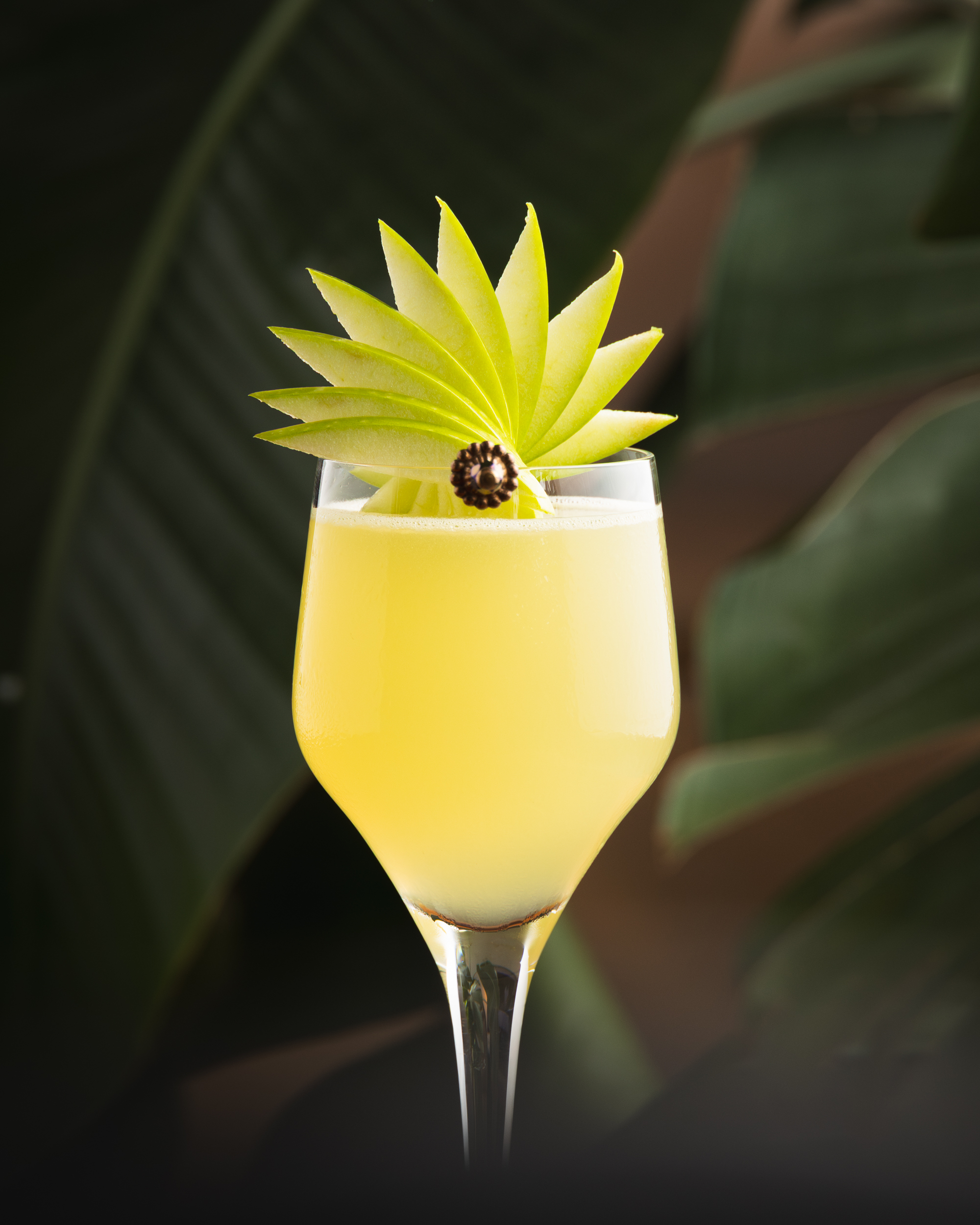 Yellow citrus cocktail in wine glass with layered green apple fan garnish, tropical foliage behind