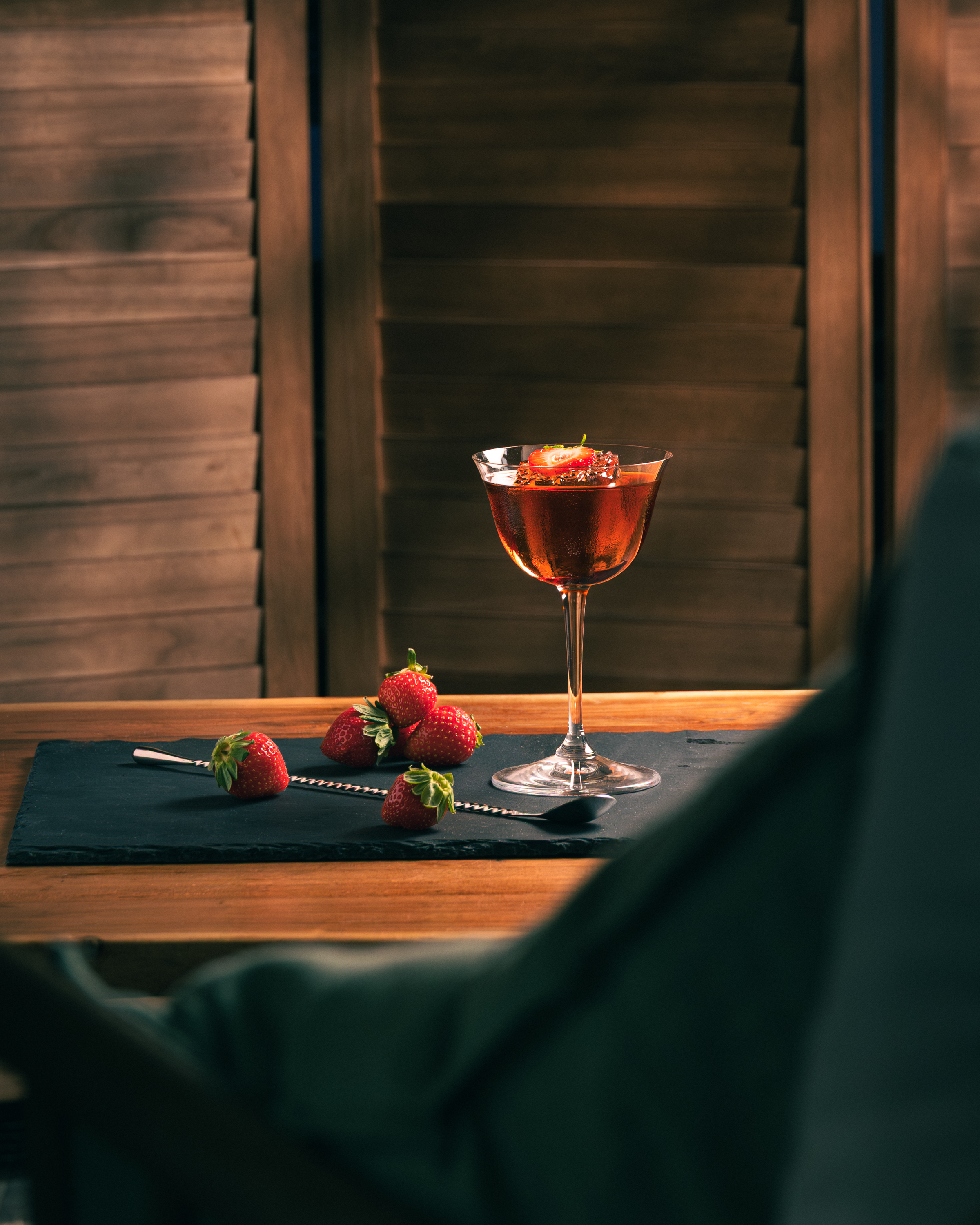 Strawberry negroni in coupe with fresh strawberries and bar spoon on slate, wood shutters behind
