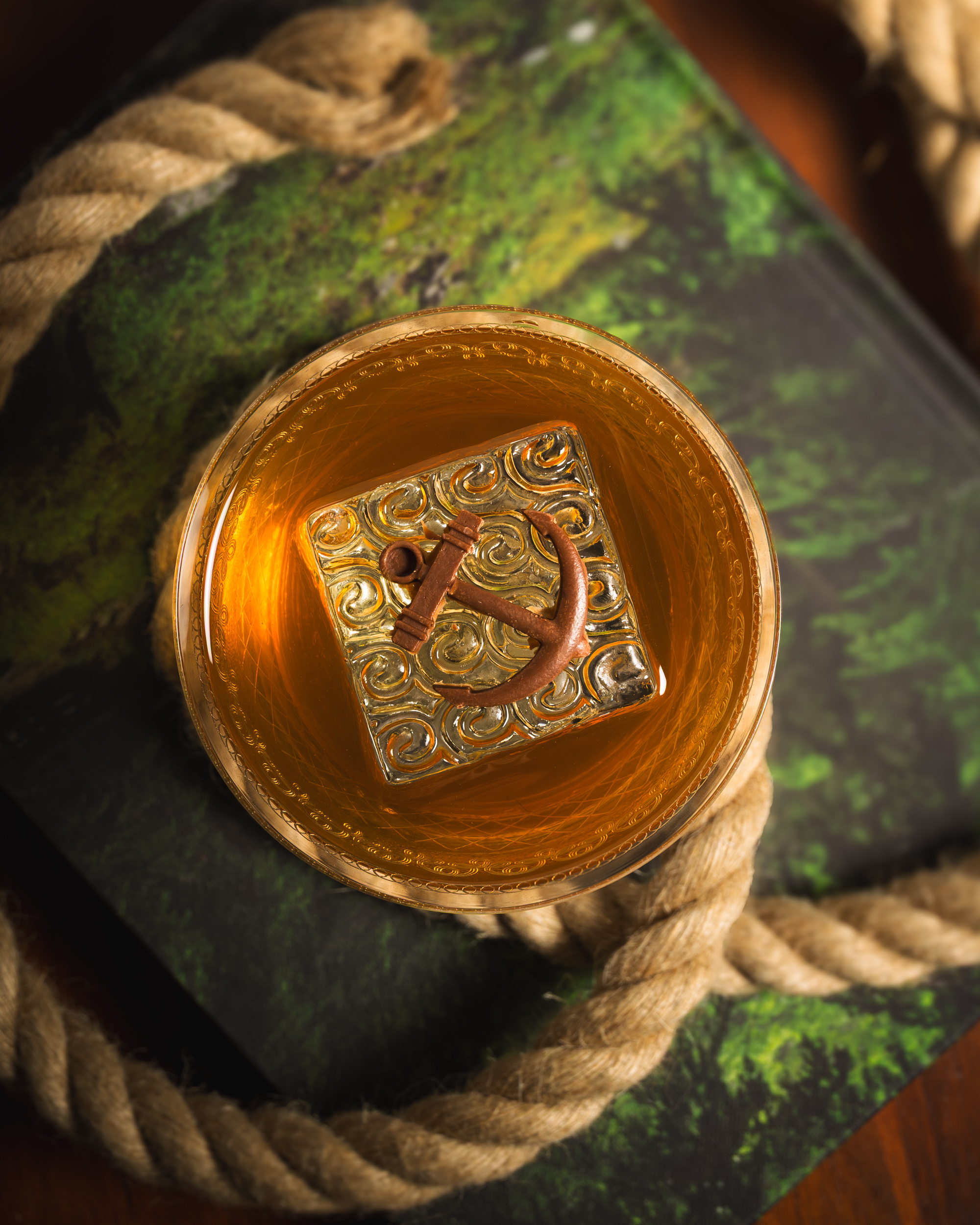 Top-down whisky old fashioned with carved anchor ice cube on a moss-covered book with rope