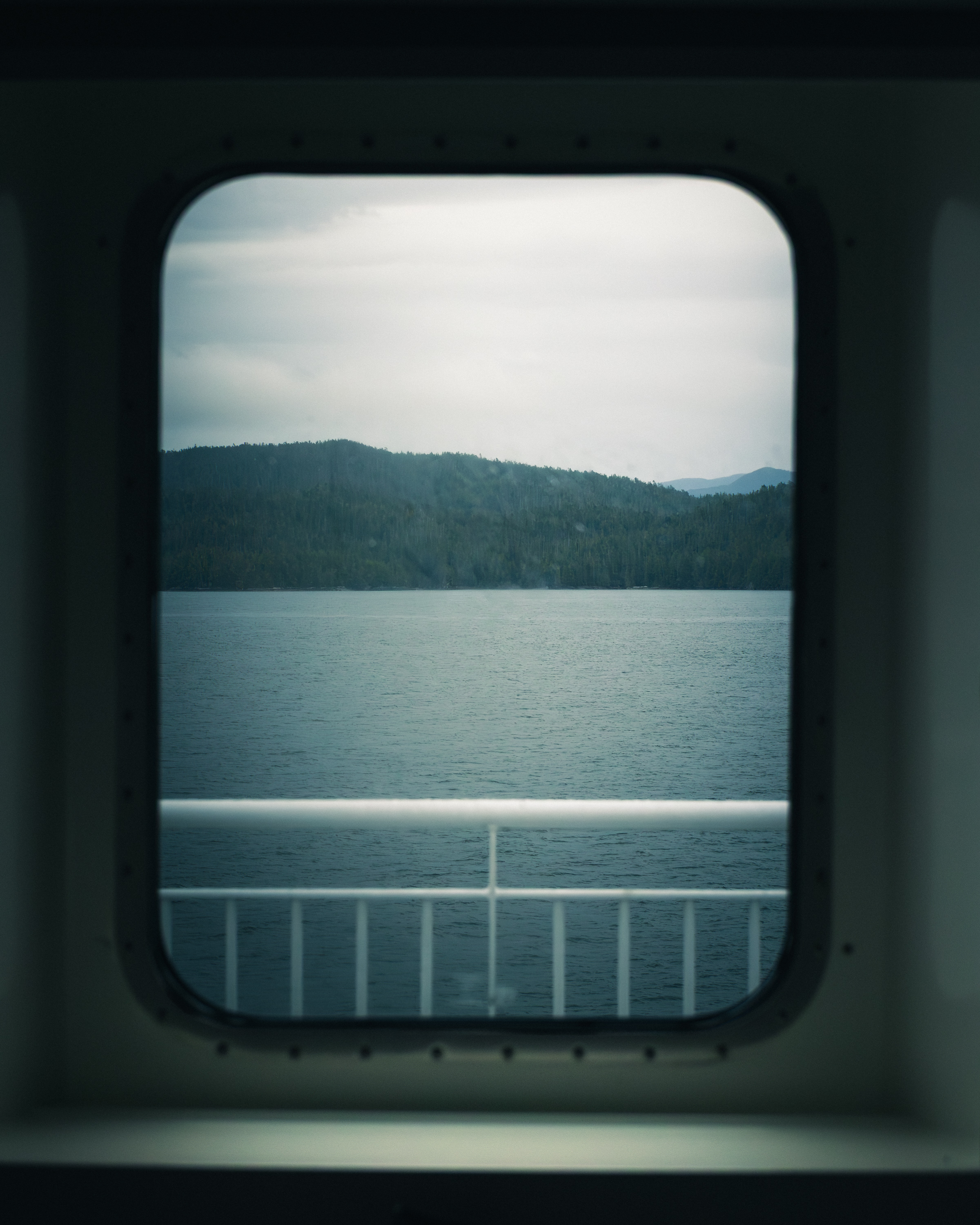 BC coast viewed through a ferry porthole window, grey water and forested hills