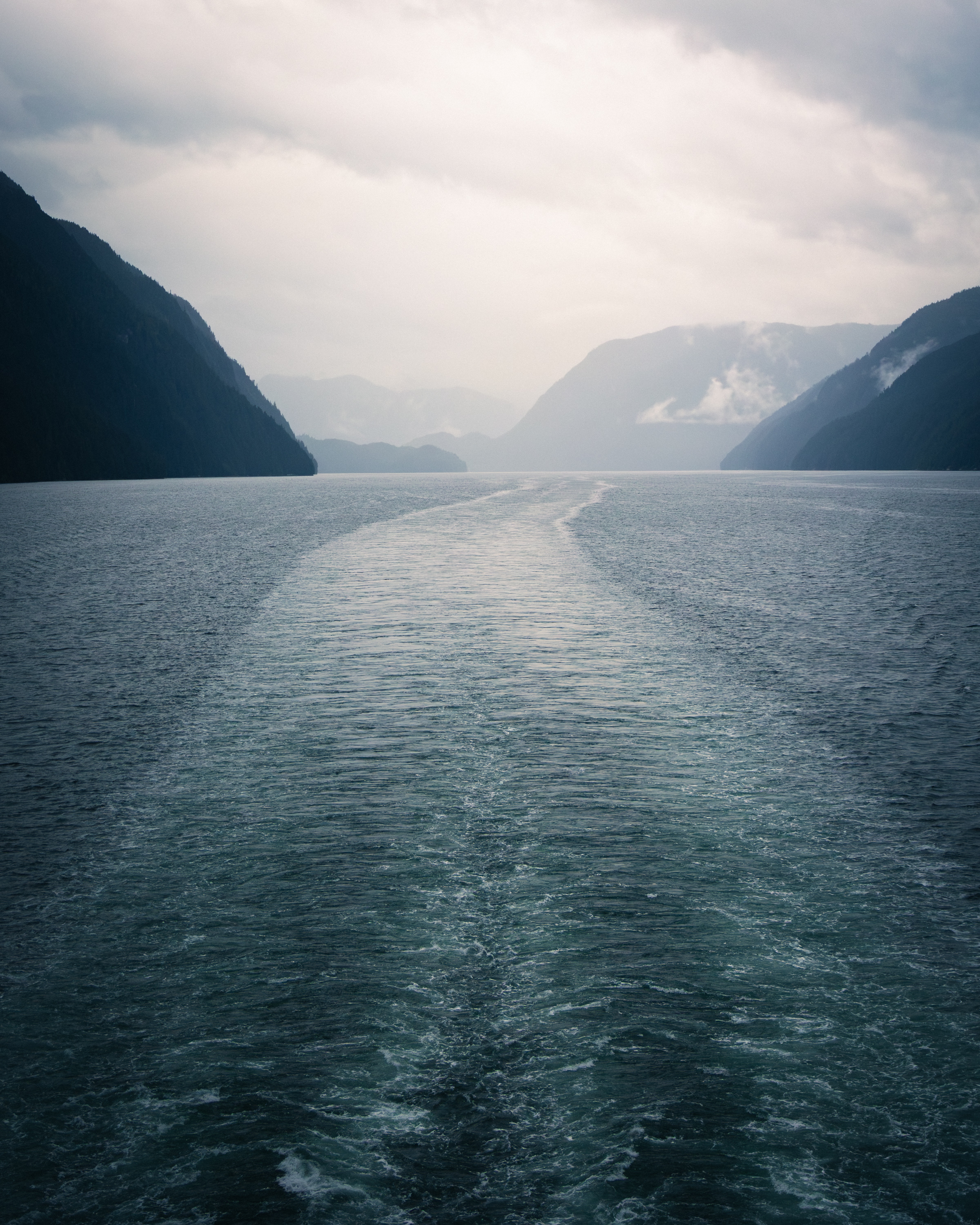 Ferry wake cutting through a narrow BC fjord flanked by dark mountain ridges