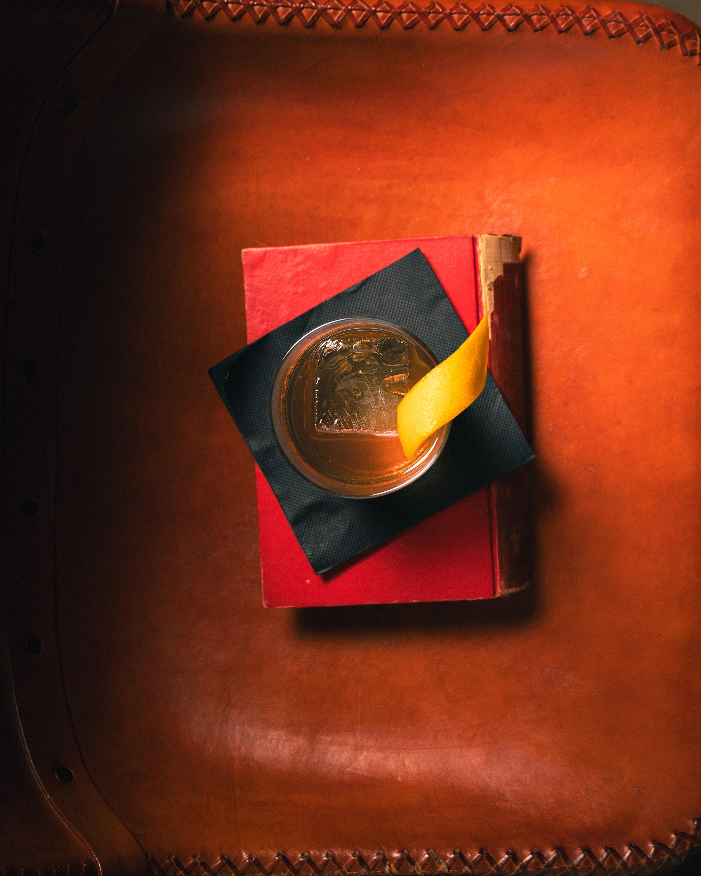 Top-down old fashioned with lemon twist resting on a red book atop a leather armchair