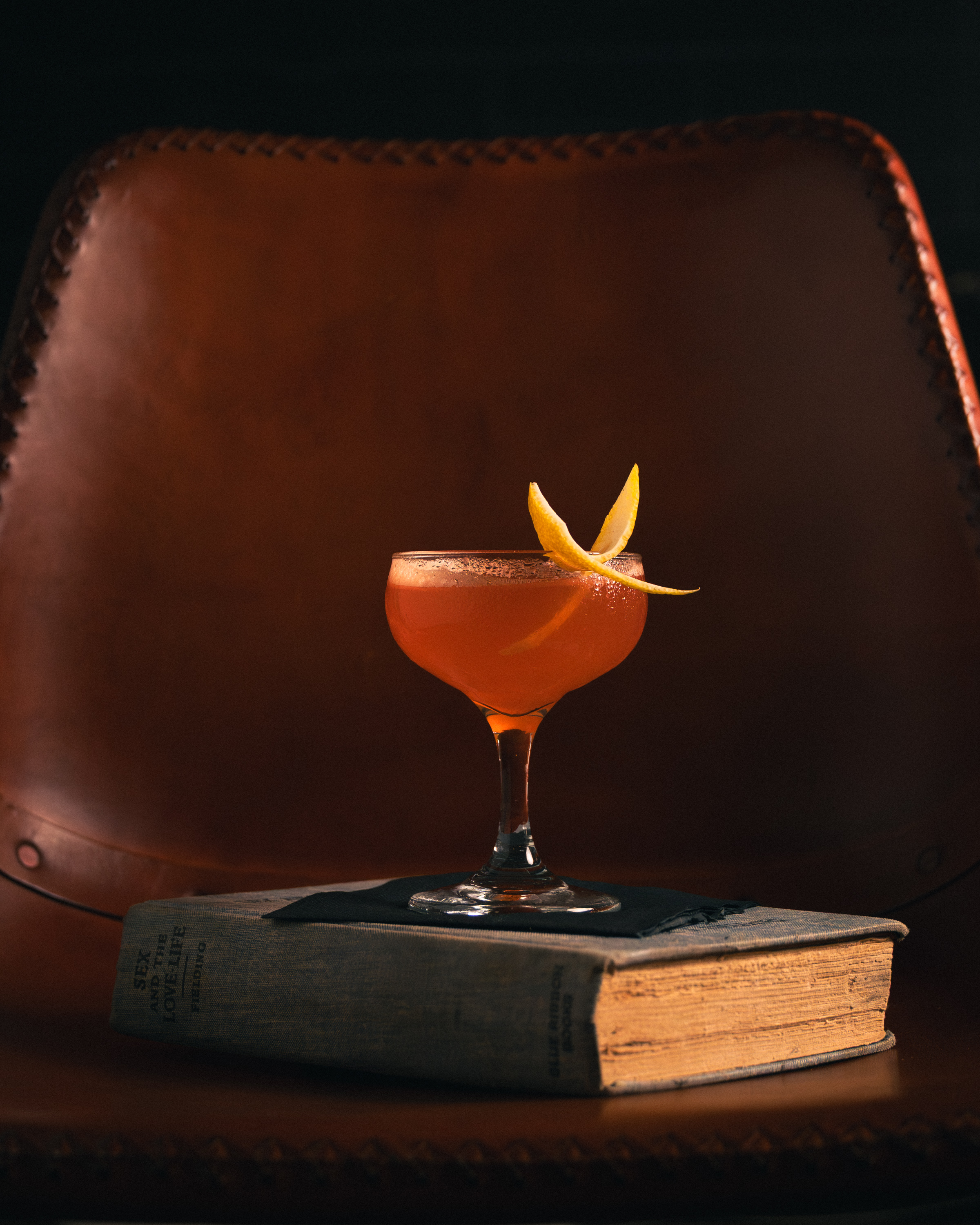 Amber cocktail in coupe with double lemon twist on a worn book, leather chair behind