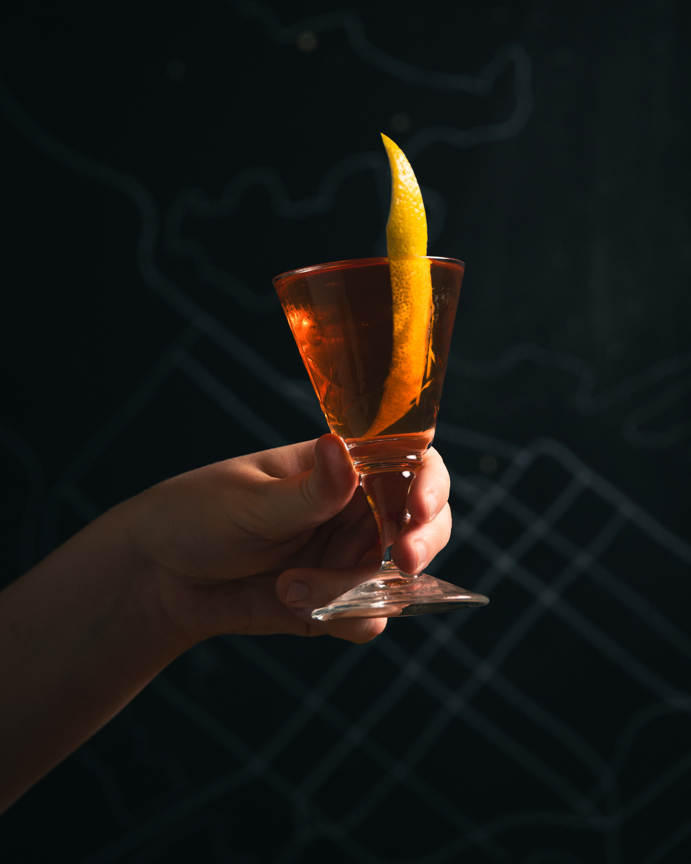 Hand holding amber cocktail with lemon twist against a dark chalk-map background