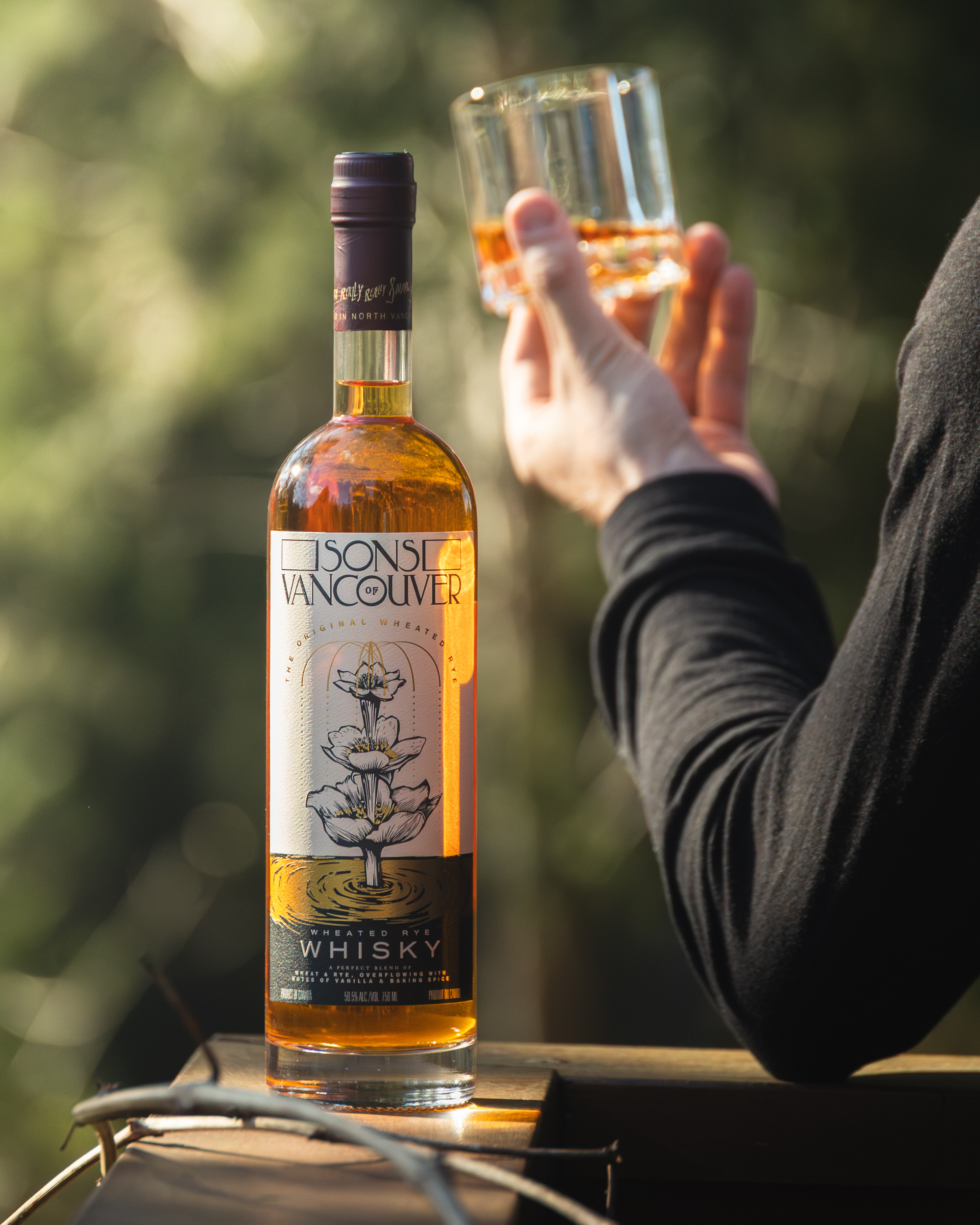 Sons of Vancouver Wheated Rye bottle outdoors with a hand holding whisky glass in forest light