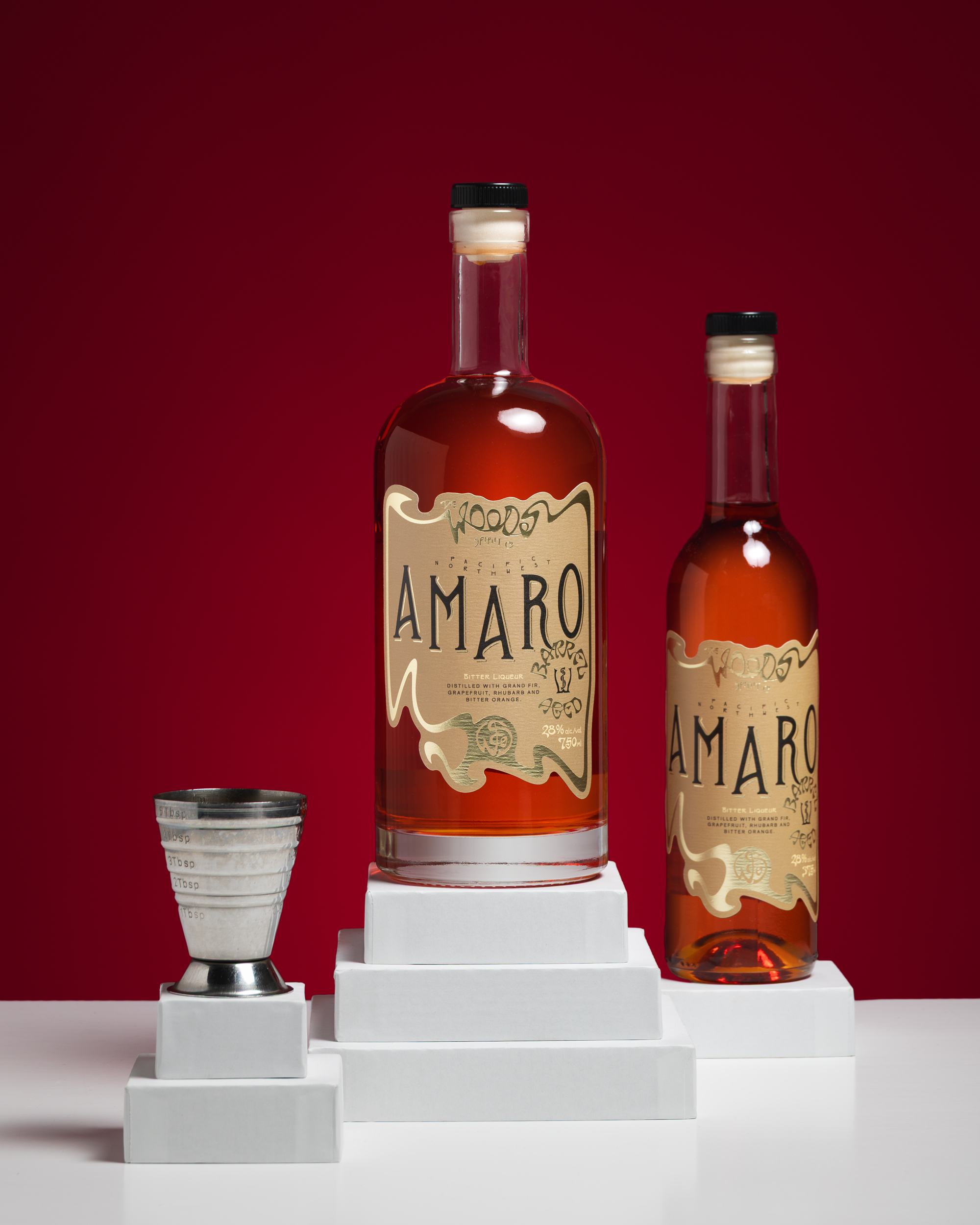Two sizes of Wood Spirit Co Barrel Aged Amaro bottles with a steel jigger on a red ground