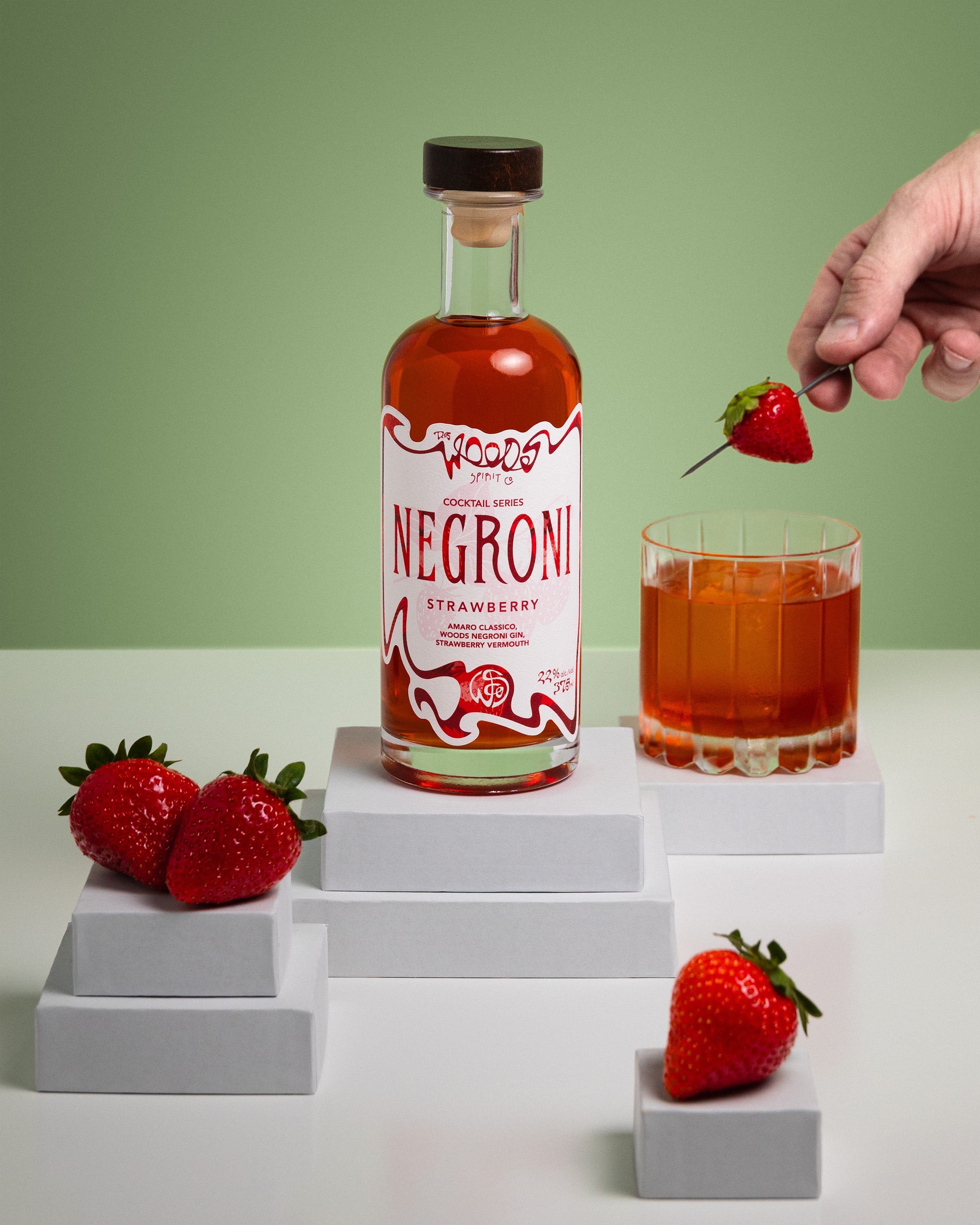 Wood Spirit Co Strawberry Negroni bottle with cocktail and hand placing strawberry garnish